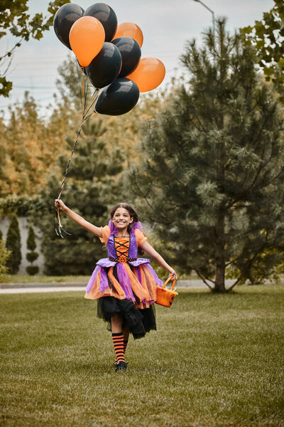 joyous girl in Halloween costume holding balloons and candy bucket while running on green grass
