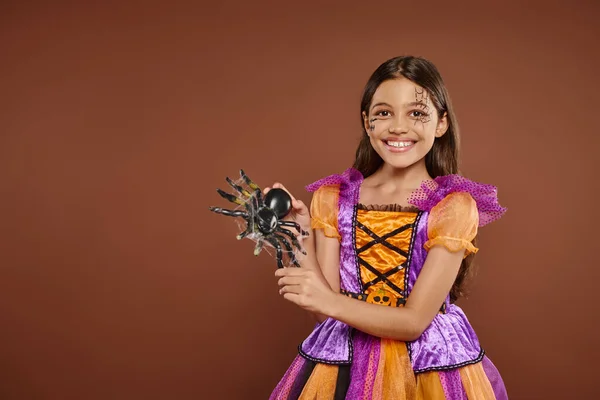 stock image joyous girl in Halloween costume with spiderweb makeup holding fake spider on brown backdrop, spooky