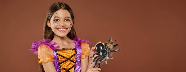 stock image joyous girl in Halloween costume with spiderweb makeup holding fake spider on brown backdrop, banner