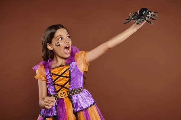 stock image scared girl in Halloween costume looking at black spider and screaming on brown backdrop, October 31
