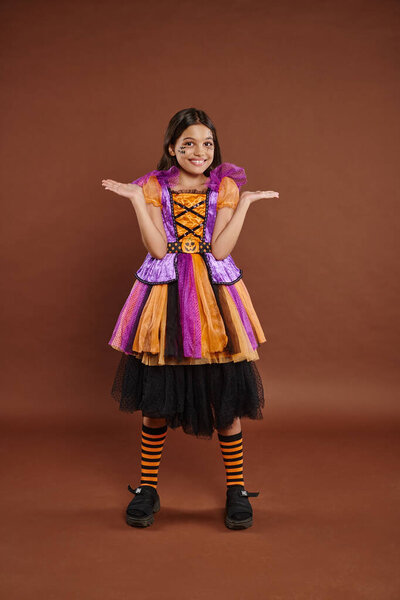 funny girl in Halloween costume with spiderweb makeup smiling and gesturing on brown backdrop