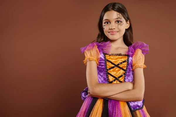 stock image pleased girl in Halloween costume with spiderweb makeup standing with folded arms on brown backdrop