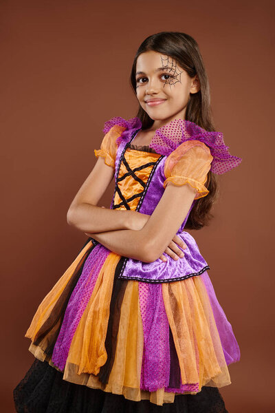happy kid in Halloween costume with spiderweb makeup standing with folded arms on brown backdrop