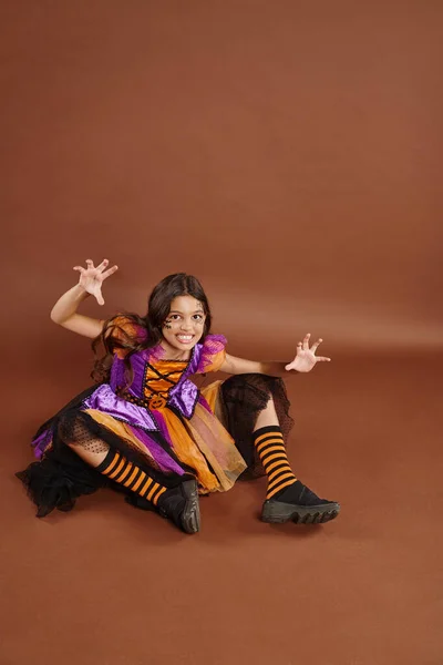 stock image spooky girl in Halloween witch costume sitting and growling on brown backdrop, October 31