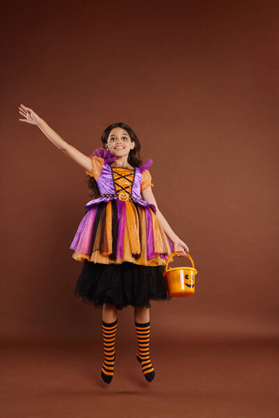 happy girl in Halloween costume levitating with candy bucket on brown background, magic concept