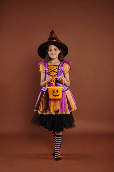 happy girl in Halloween costume and pointed hat standing with candy bucket on brown background