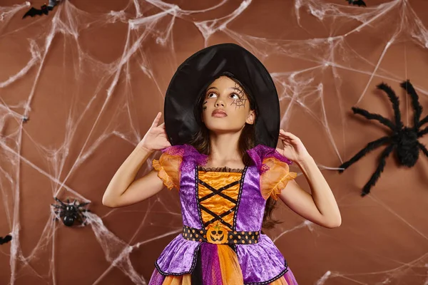 身着万圣节服装头戴女巫帽的女孩抬起头 站在褐色背景的蜘蛛网旁边 — 图库照片