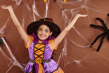 joyous little witch in Halloween costume and pointed hat with raised hands on brown backdrop clipart