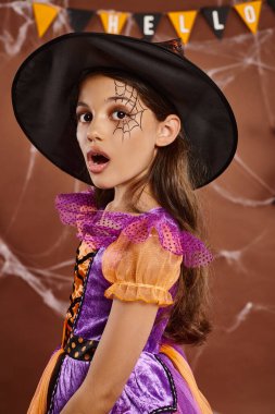 shocked little witch in Halloween costume and pointed hat on brown backdrop, surprised girl clipart