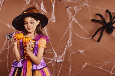 cheerful girl in Halloween witch costume and pointed hat holding pumpkins on brown backdrop clipart