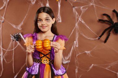 cheerful girl with spiderweb makeup in Halloween costume holding pumpkins on brown backdrop clipart