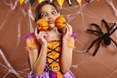 Örümcek ağı makyajlı komik kız yanakları şişiriyor ve kahverengi arka planda balkabağı tutuyor, Cadılar Bayramı