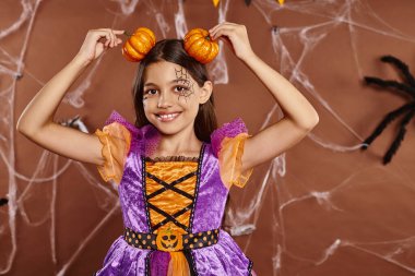funny girl with spiderweb makeup smiling and holding pumpkins near head on brown backdrop, Halloween clipart