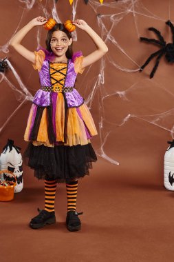 cheerful girl in Halloween bright dress holding pumpkins near head on brown backdrop, spooky season clipart