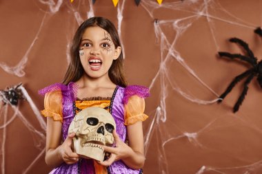 Cadılar Bayramı kostümlü kız kafatası tutuyor ve kahverengi arka planda kameraya hırlıyor. Ürkütücü sezon.
