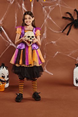 cheerful girl in dress holding skull and smiling on brown background, Halloween spooky season clipart