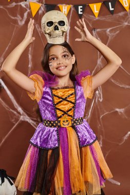 positive girl in Halloween costume standing with skull on head on brown background, spooky season clipart
