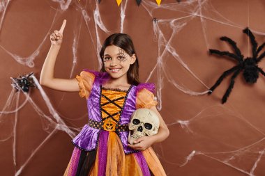 Cadılar Bayramı kıyafetli neşeli kız kafatası ile ayakta duruyor ve kahverengi arka planda parmağını kaldırıyor.