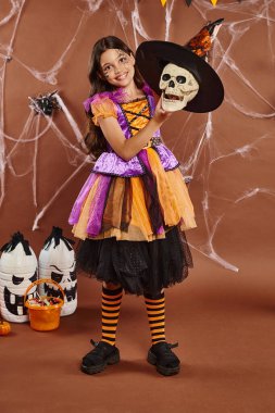 Cadılar Bayramı kostümlü mutlu kız kahverengi arka planda cadı şapkası takıyor, ürkütücü sezon.