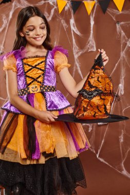 cheerful kid in Halloween witch costume holding pointed hat on brown background, spooky season clipart
