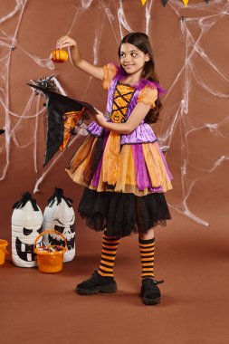 happy child in Halloween witch costume holding pumpkin near pointed hat on brown background clipart