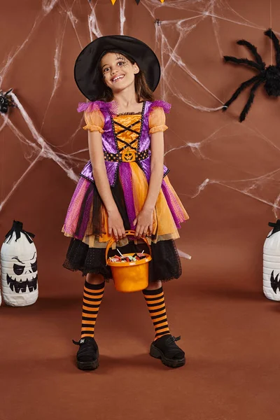 happy girl in witch hat and Halloween costume holding bucket with sweets near diy spooky decor