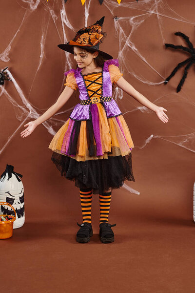 positive girl in witch costume and pointed hat gesturing near sweets bucket, Halloween concept
