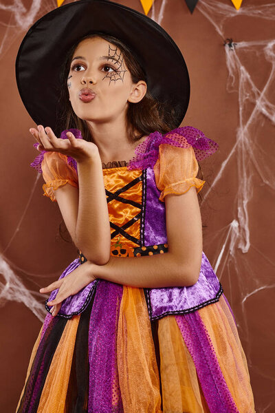 cute girl in witch costume and pointed hat sending air kiss on brown backdrop, Halloween concept