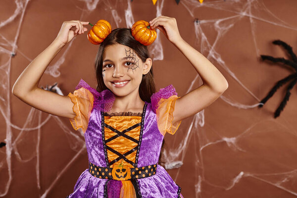 funny girl with spiderweb makeup smiling and holding pumpkins near head on brown backdrop, Halloween