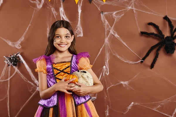 stock image cheerful girl in Halloween costume holding pumpkins and skull on brown backdrop, spooky season