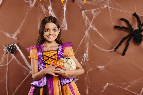 cheerful girl in Halloween costume holding pumpkins and skull on brown backdrop, spooky season