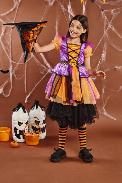 cheerful child in Halloween witch costume holding pointed hat upside down on brown background