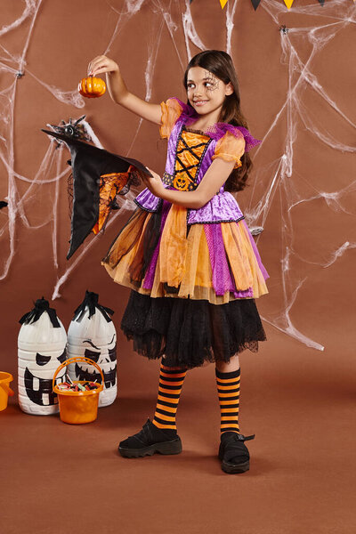 happy child in Halloween witch costume holding pumpkin near pointed hat on brown background