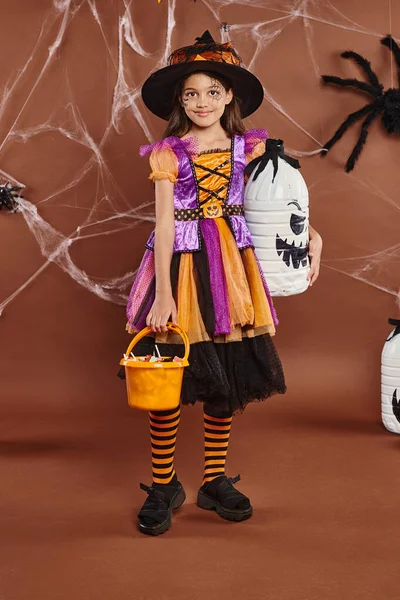 happy girl in witch hat and Halloween costume holding bucket with sweets and diy spooky decor