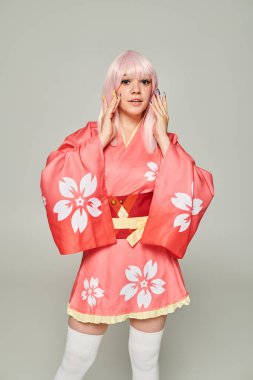 Renkli manikürlü genç sarışın kadın pembe kimono giyiyor ve gri kameraya bakıyor.
