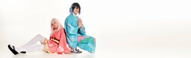 Kimonolu bir adam, dua eden elleriyle anime kadının yanında beyaz, yatay pankartta oturan sarı peruklu bir kadın.