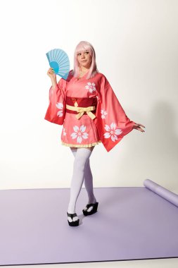Anime stili kadın pembe kimonolu ve sarı peruklu. Beyaz stüdyoda mor halıda yelpaze.