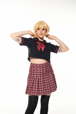 stylish tattooed anime woman in school uniform touching ponytails on yellow blonde wig on white clipart