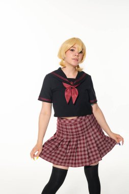 Sarı peruklu, ekose etekli karınca pozu veren genç anime tarzı kadın beyaz kameraya bakıyor.