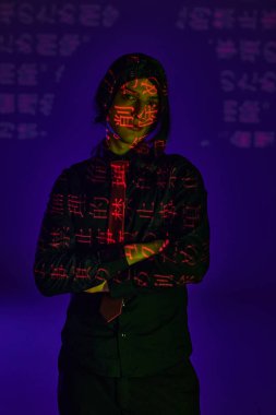 student in black wig and uniform with folded arms in neon light with hieroglyphs on blue backdrop clipart