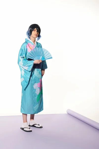 stock image young man in blue kimono and wig posing with hand fan on purple carpet and white backdrop, cosplay