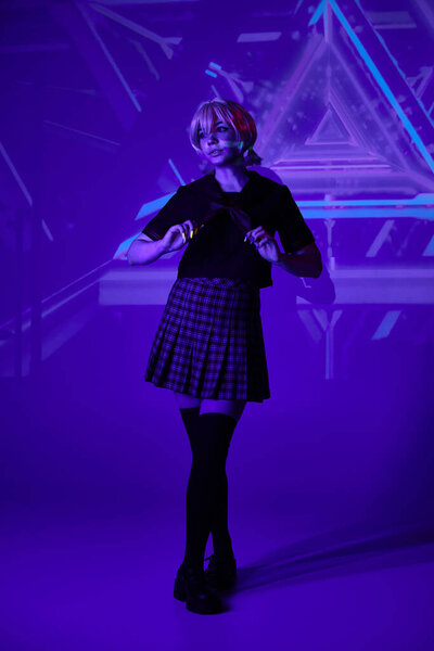 anime woman in blonde wig and school uniform in blue neon light on abstract backdrop, full length