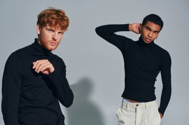two multiracial male models with accessories in black turtlenecks gesturing and looking a camera clipart