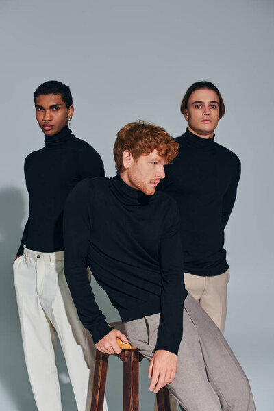 red haired bearded man in turtleneck sitting on chair with other two men behind him, gray backdrop