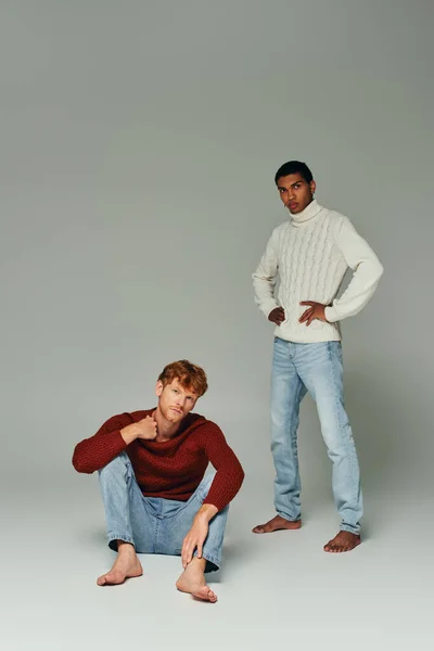 stock image red haired man in sweater squatting on floor with african american man standing on gray background