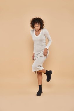 cheerful african american woman walking in midi dress and boots on beige backdrop, autumn fashion clipart