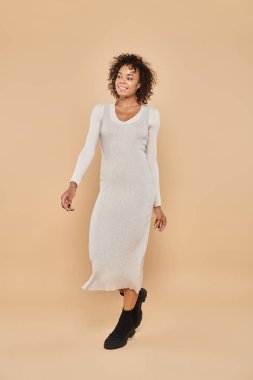 brunette african american woman standing in midi dress and boots on beige backdrop, autumn fashion clipart