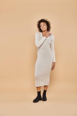 happy african american woman  in midi dress and boots adjusting curly hair on beige backdrop clipart