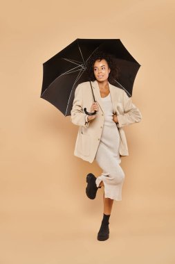 Midi elbisesi ve bej üzerinde şemsiye altında duran sonbahar ceketli şık Afrikalı Amerikalı kadın.