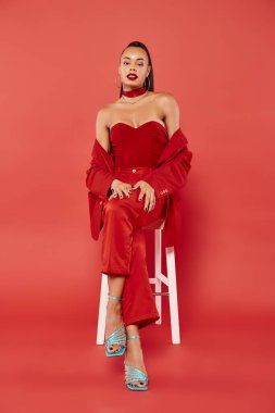 Uzun boylu, Afro-Amerikan kadın takım elbiseli ve kırmızı sandalyede oturan yüksek topuklu.